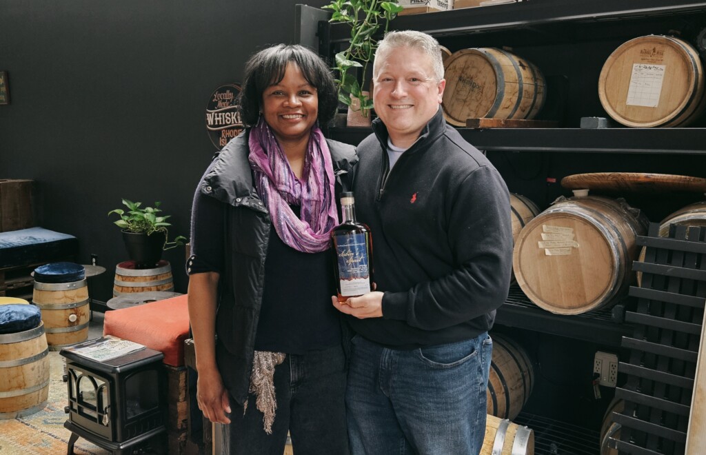 A woman and man hold up a whiskey bottle in front of a display of aging oak barrels.