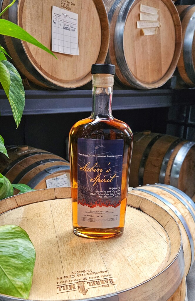 A bottle of whiskey sitting on an oak barrel in a tasting room. The label is blue and has a flame design.