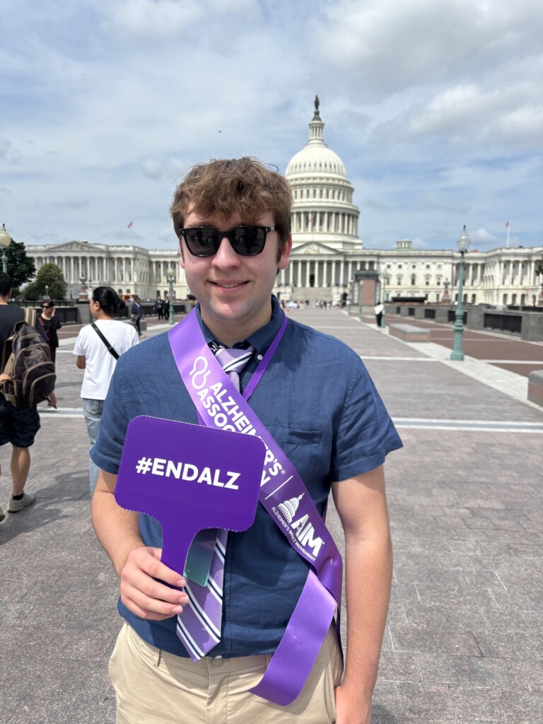 A young man in a shirt, tie and sunglasses smiles outside the Capitol building while holding a purple sign that says "ENDALZ."
