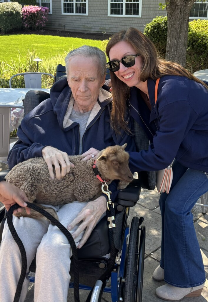An elderly man in a wheelchair pets a goat on his lap while a woman smiles next to him.