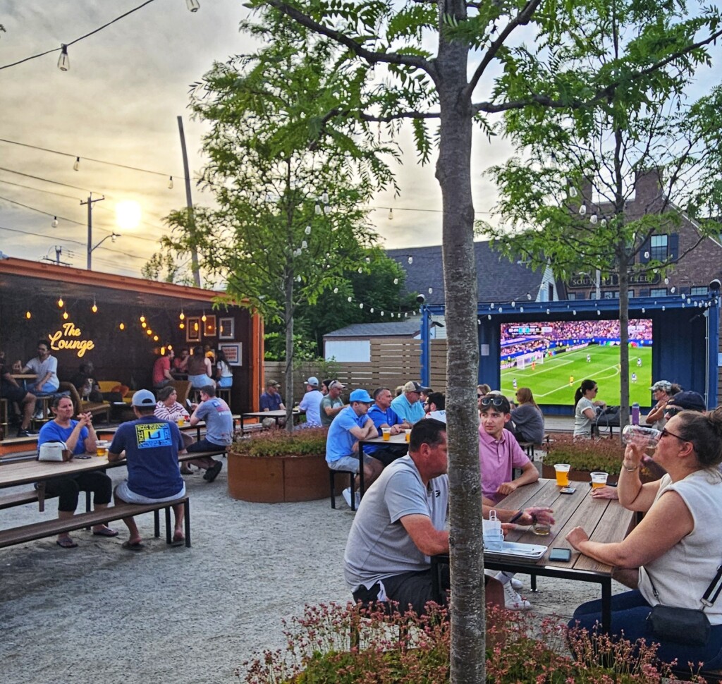 An outdoor beer garden features people sitting at picnic tables and string lights with a World Cup game on a big screen.